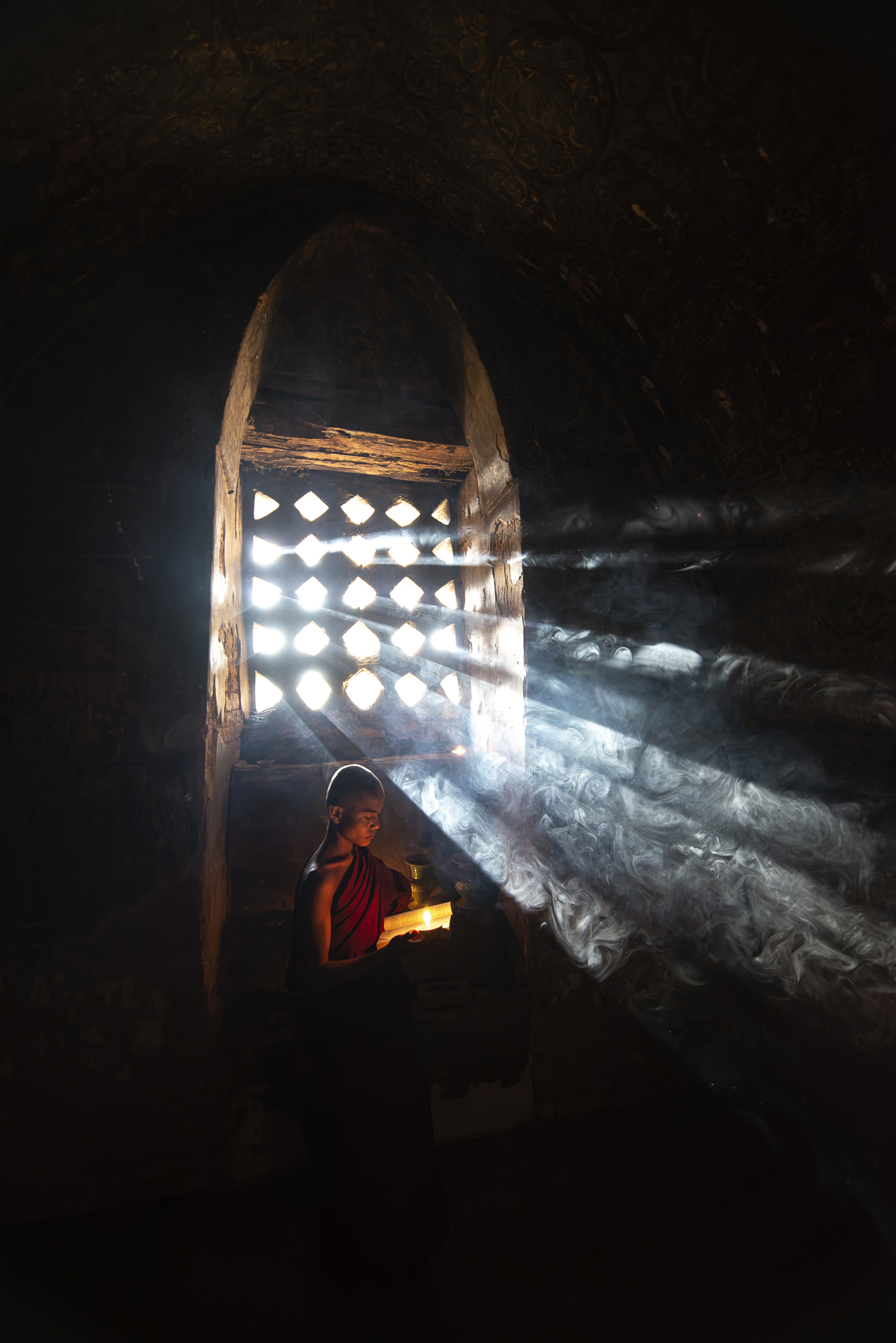 Young Buddhist monk holding a book in a sunlit temple room with dramatic light rays and incense smoke, travel photography by Gonzalo Bendito