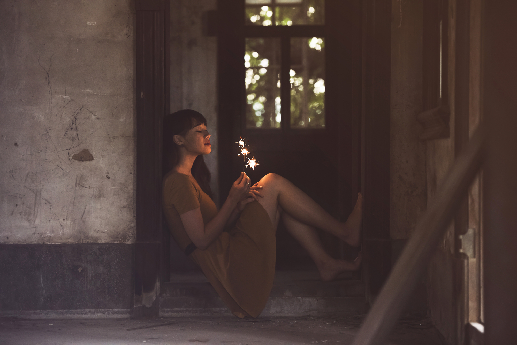 Woman in a yellow dress sitting in a rustic doorway holding glowing sparklers, portrait photography by Gonzalo Bendito