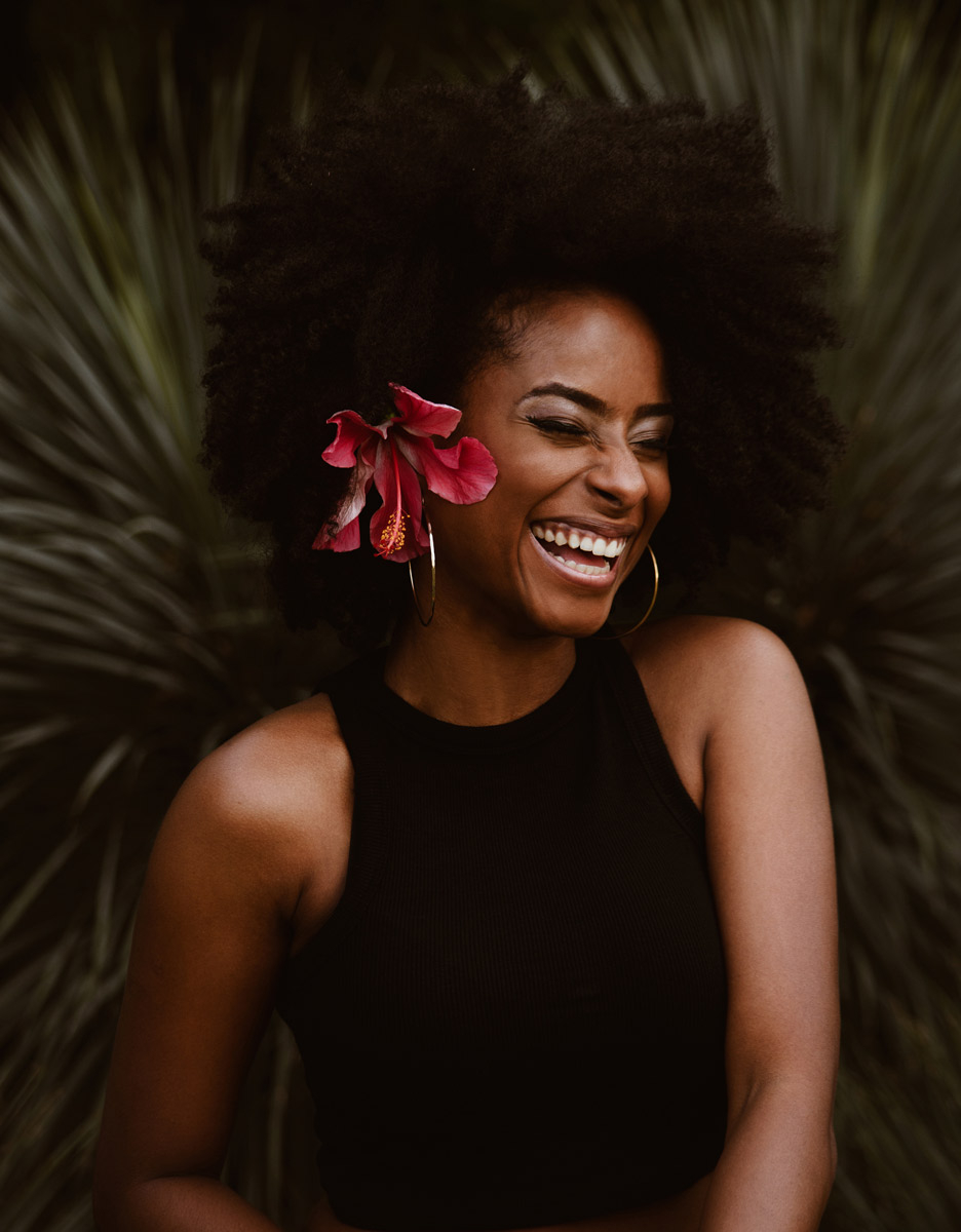 Woman laughing with a hibiscus flower tucked behind her ear against dark foliage