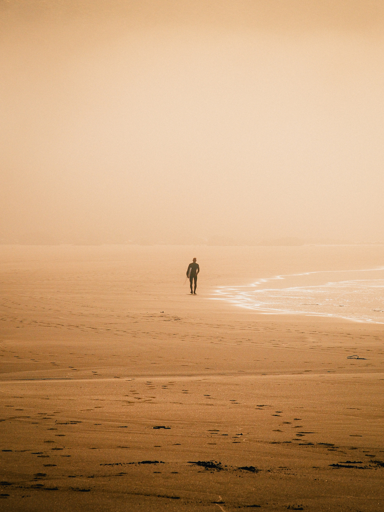Lone surfer walking across a wide sand flat in warm coastal haze