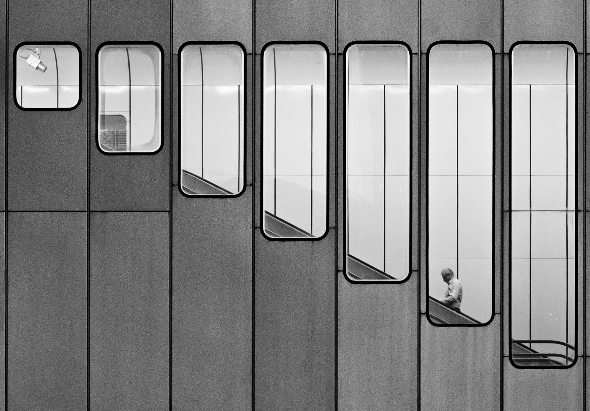 Flat-on view of a grey panel facade punctuated by tall rounded windows. A lone man descends an escalator in one of them.