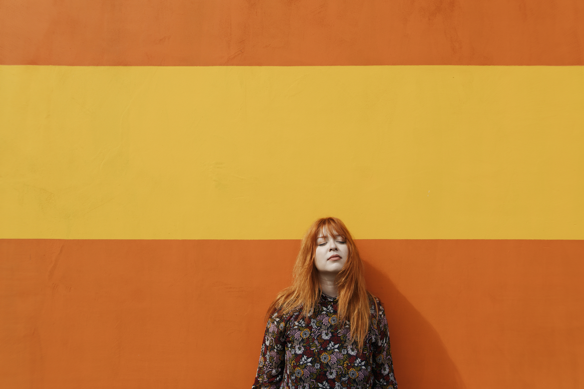 Portrait of a woman with red hair relaxing against a bold orange and yellow striped wall in an urban color photography setting.