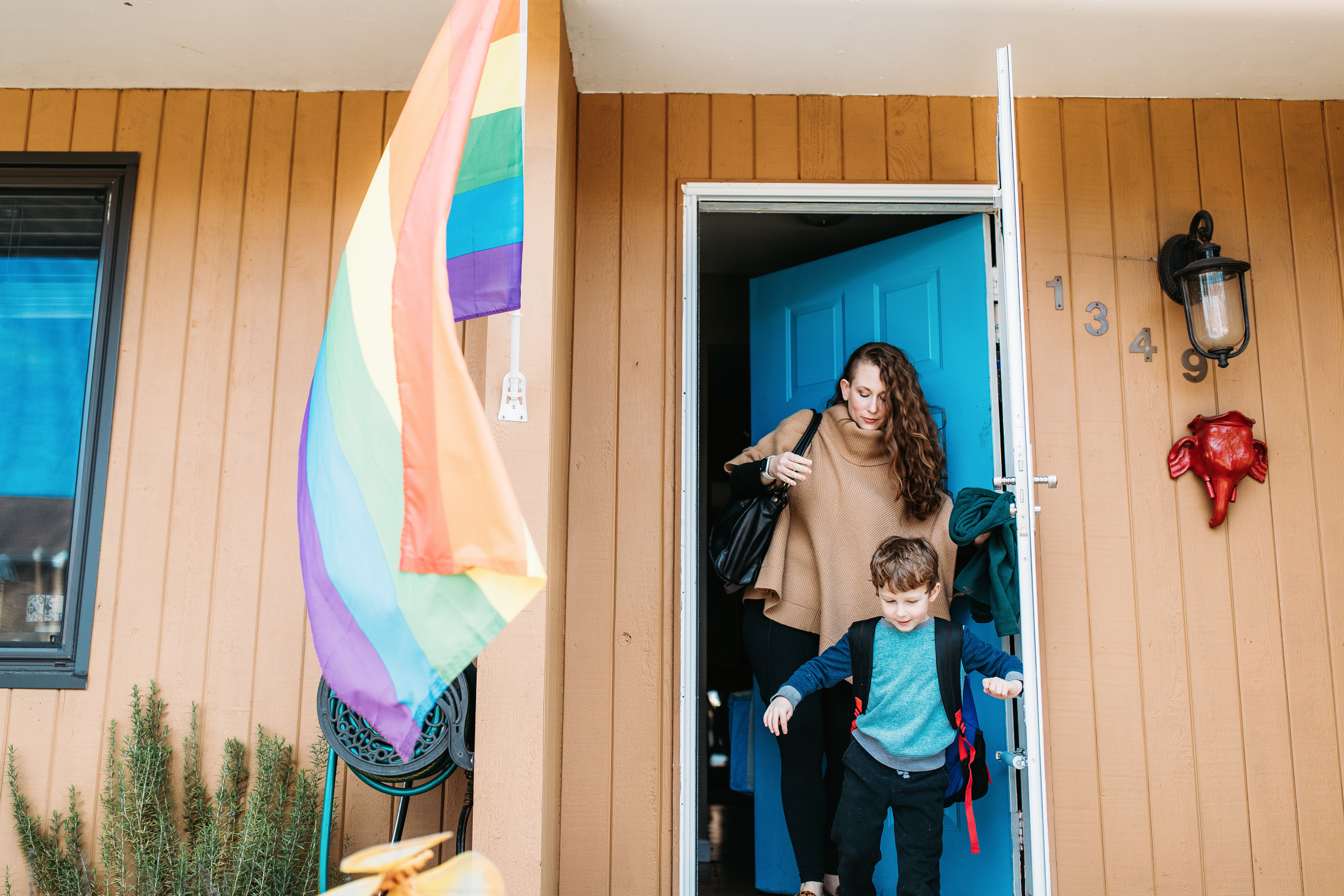 Candid photo of a woman and young boy leaving a house with a blue door, a rainbow flag waving in the foreground.