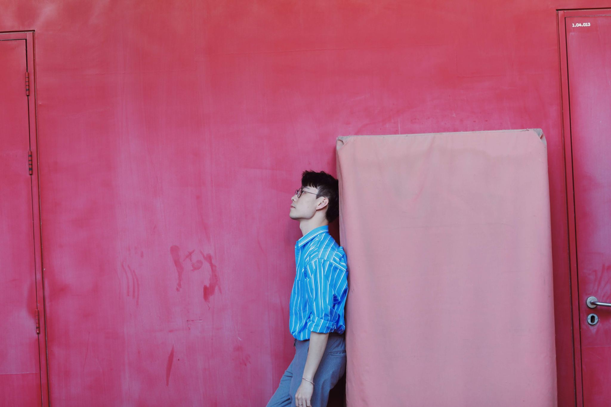 Man in a blue striped shirt resting against a pink mattress on a red wall, complementary warm and cool tones building quiet tension.