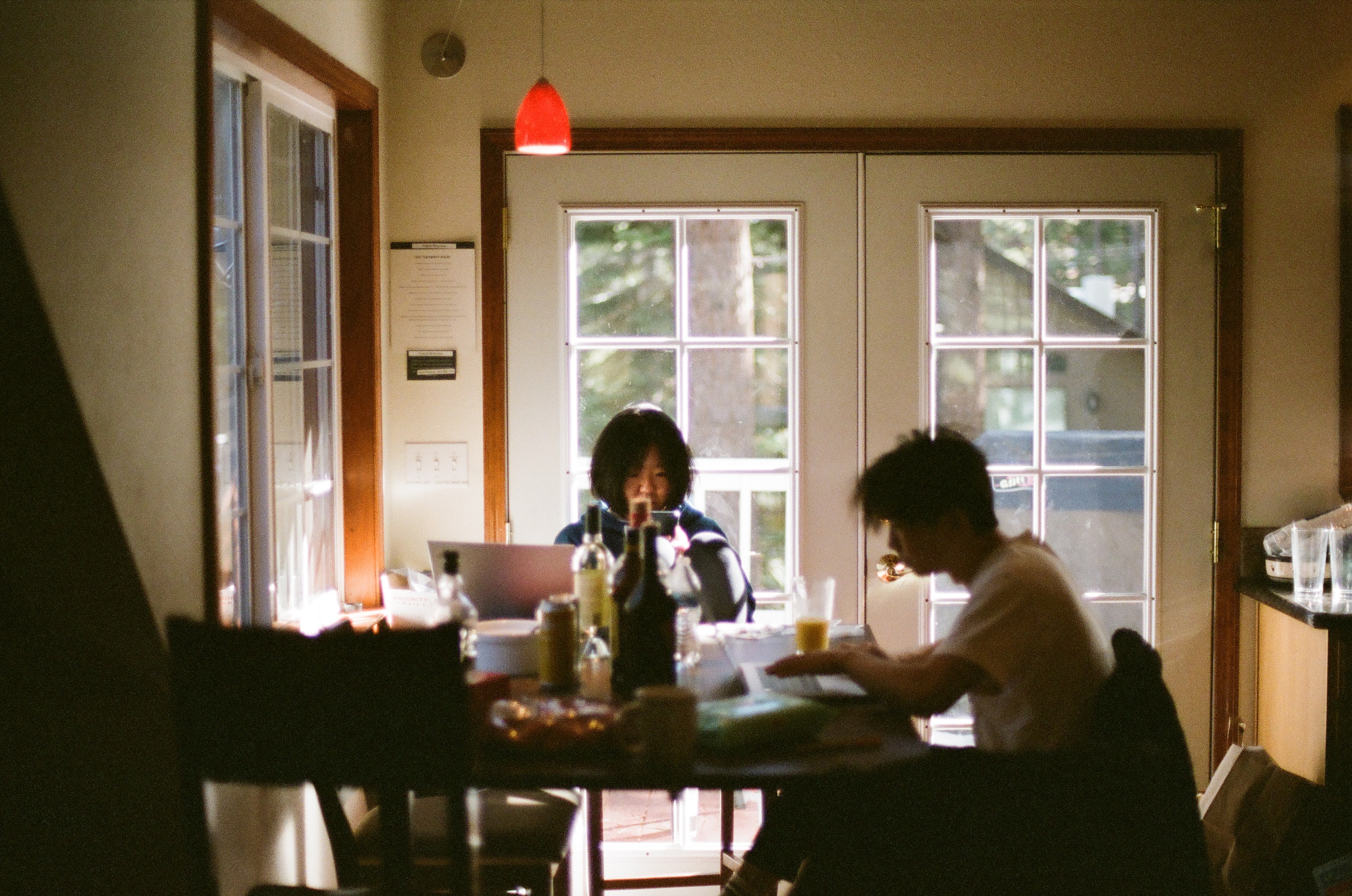 Documentary-style photo of two people working at a cluttered kitchen table, backlit by sunlight through French doors.