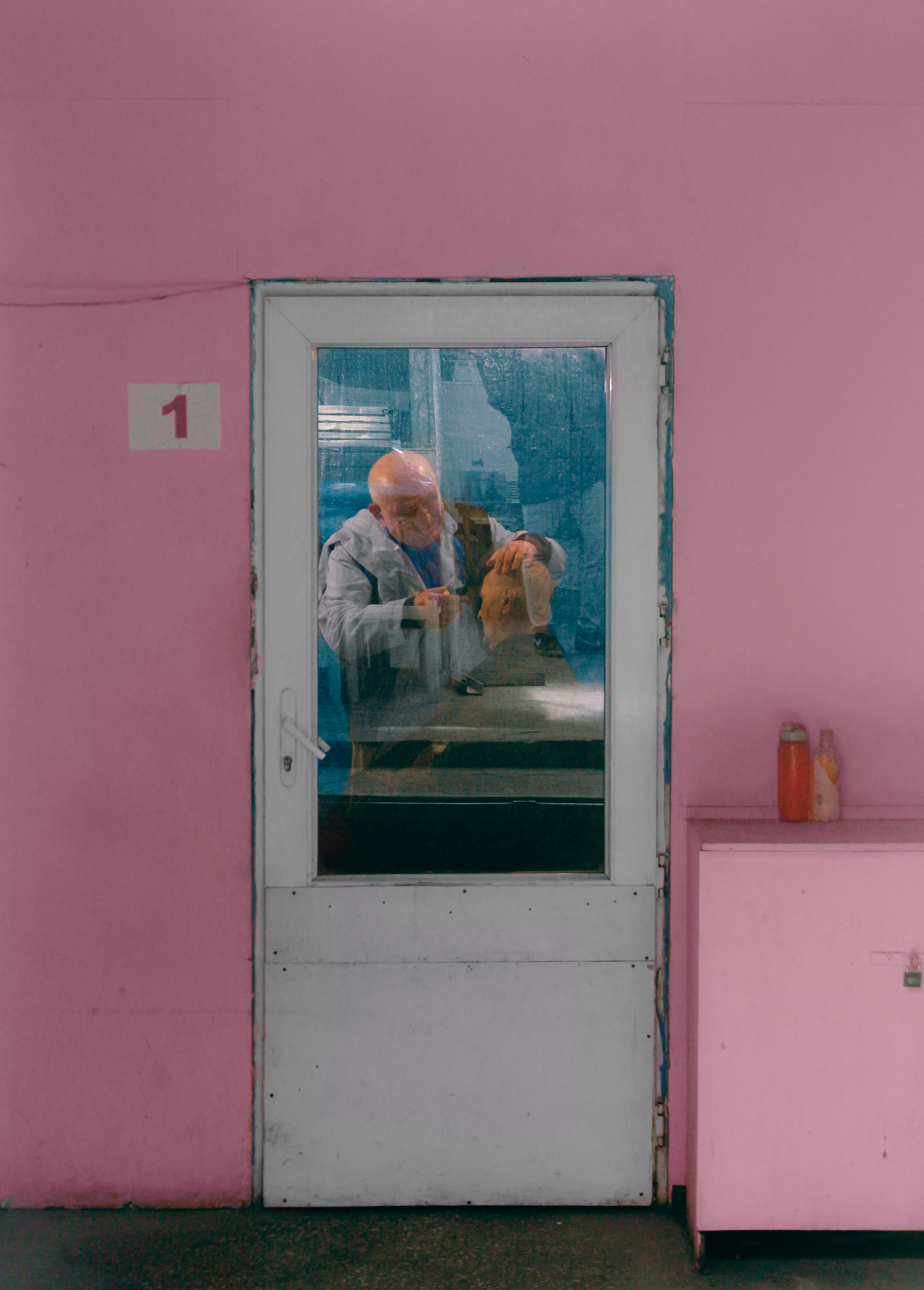 Street photography composition: through a glass door in a pink wall, an older man in a white coat works on a sculpted head.