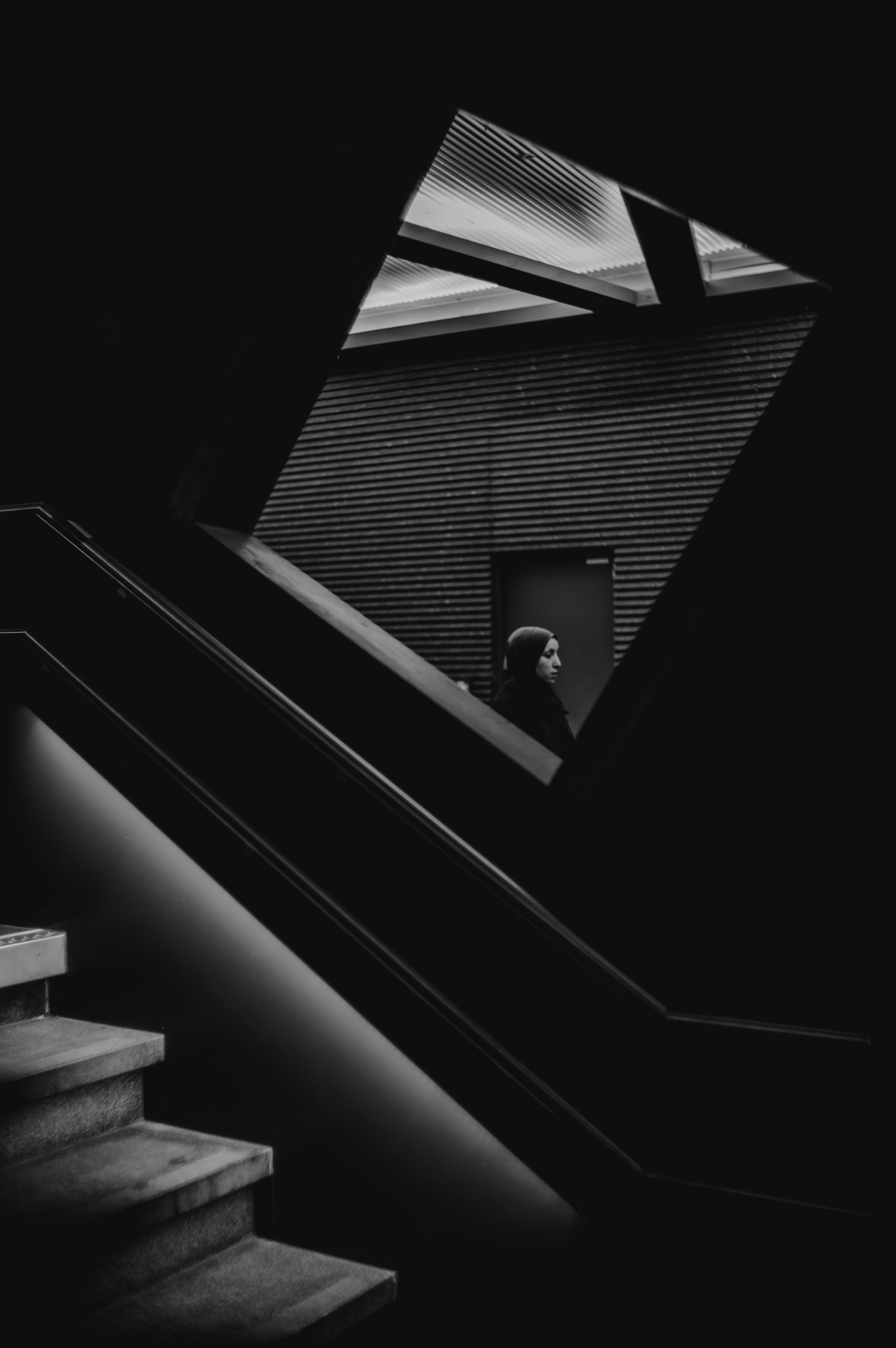 Woman in a headscarf framed in a sliver of light beyond dark stairwell geometry, an example of Eline Deckers's high-contrast street photography.