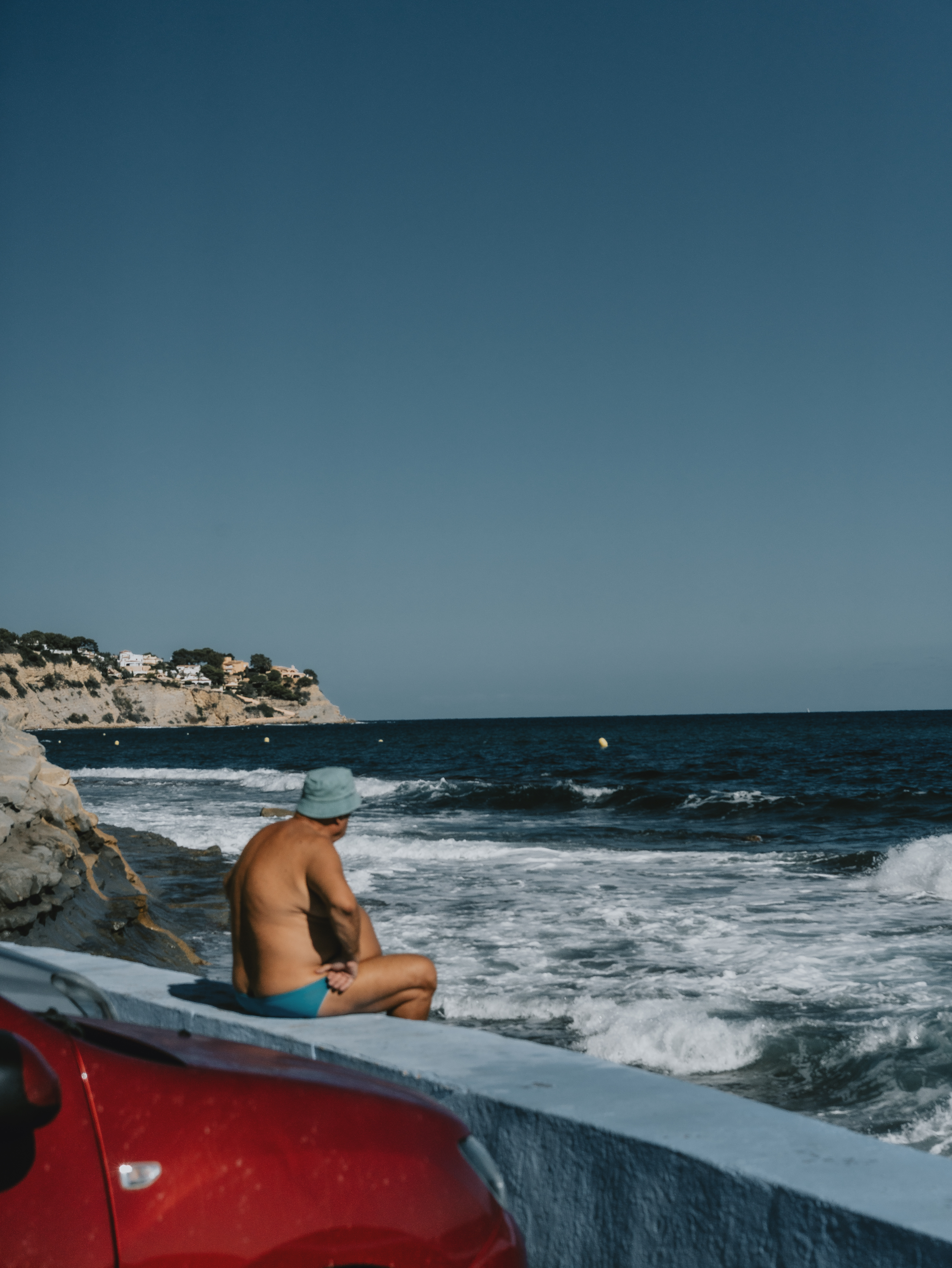 Man in a bucket hat sitting on a white sea wall watching the surf on the Costa Blanca, one of Eline Deckers's rare color frames.