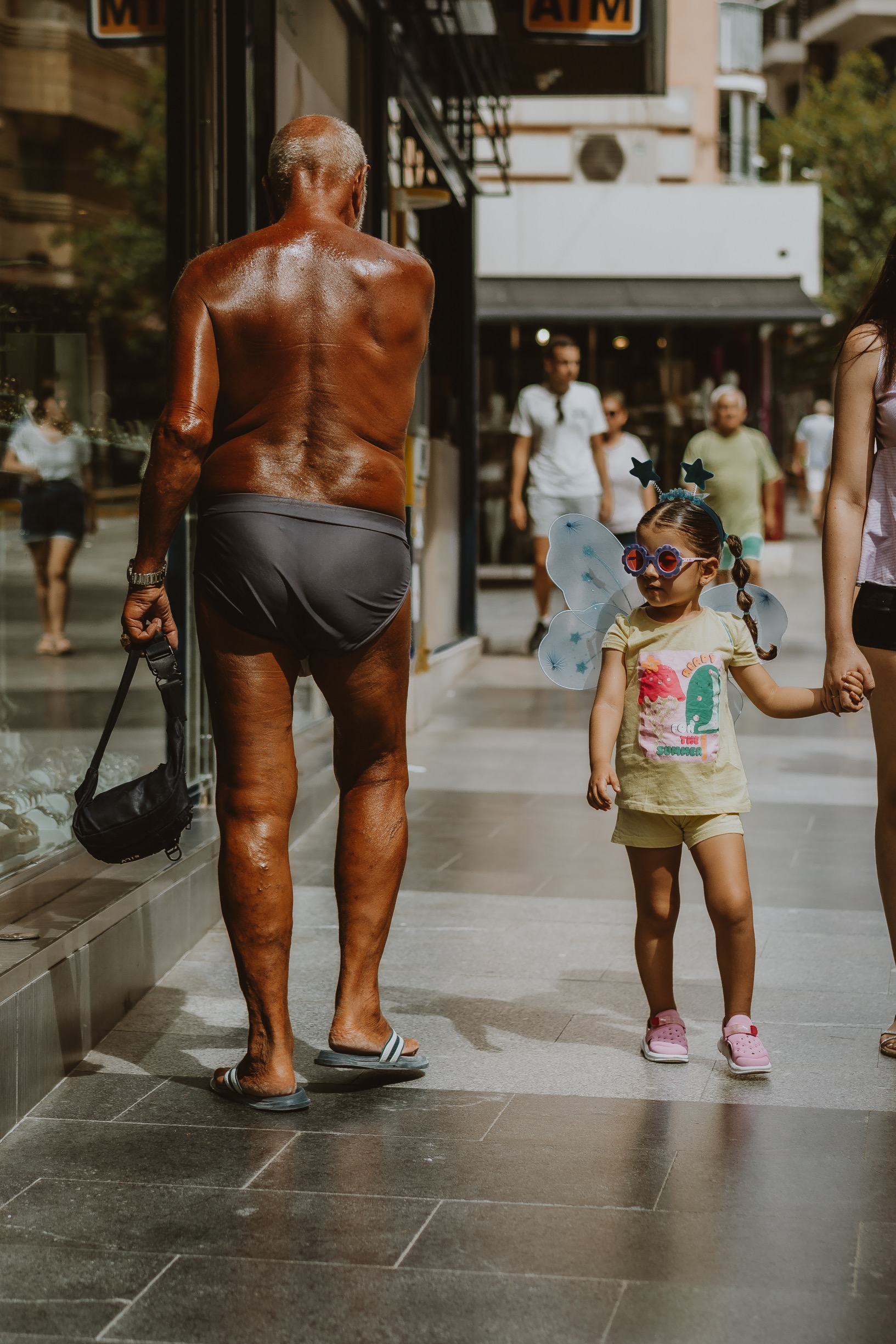 Elderly man in swim briefs walking a Spanish shopping street past a girl in fairy wings, from Eline Deckers's street photography.