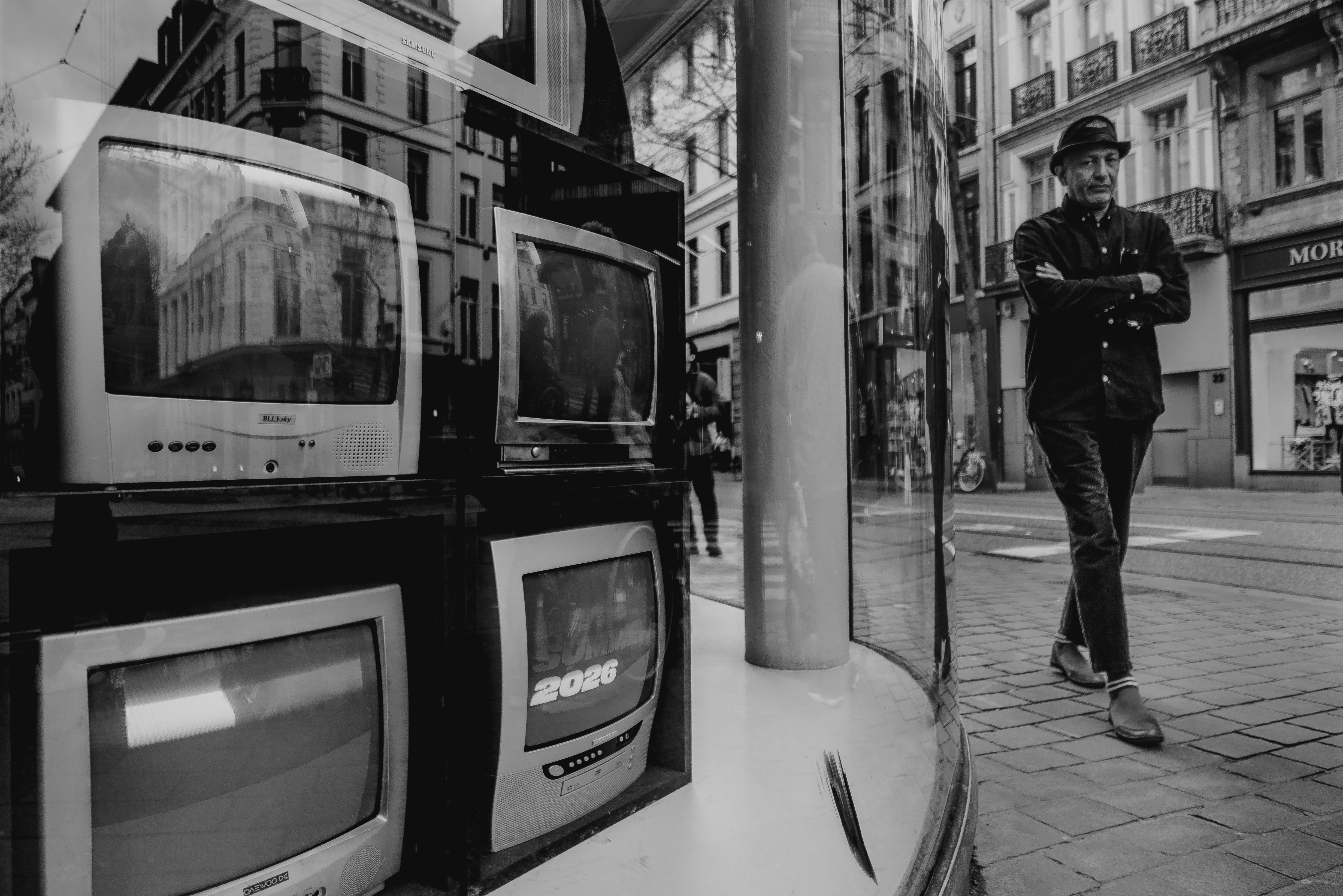 Stack of old televisions in a shop window as a man in a flat cap walks past, from Eline Deckers's street photography.