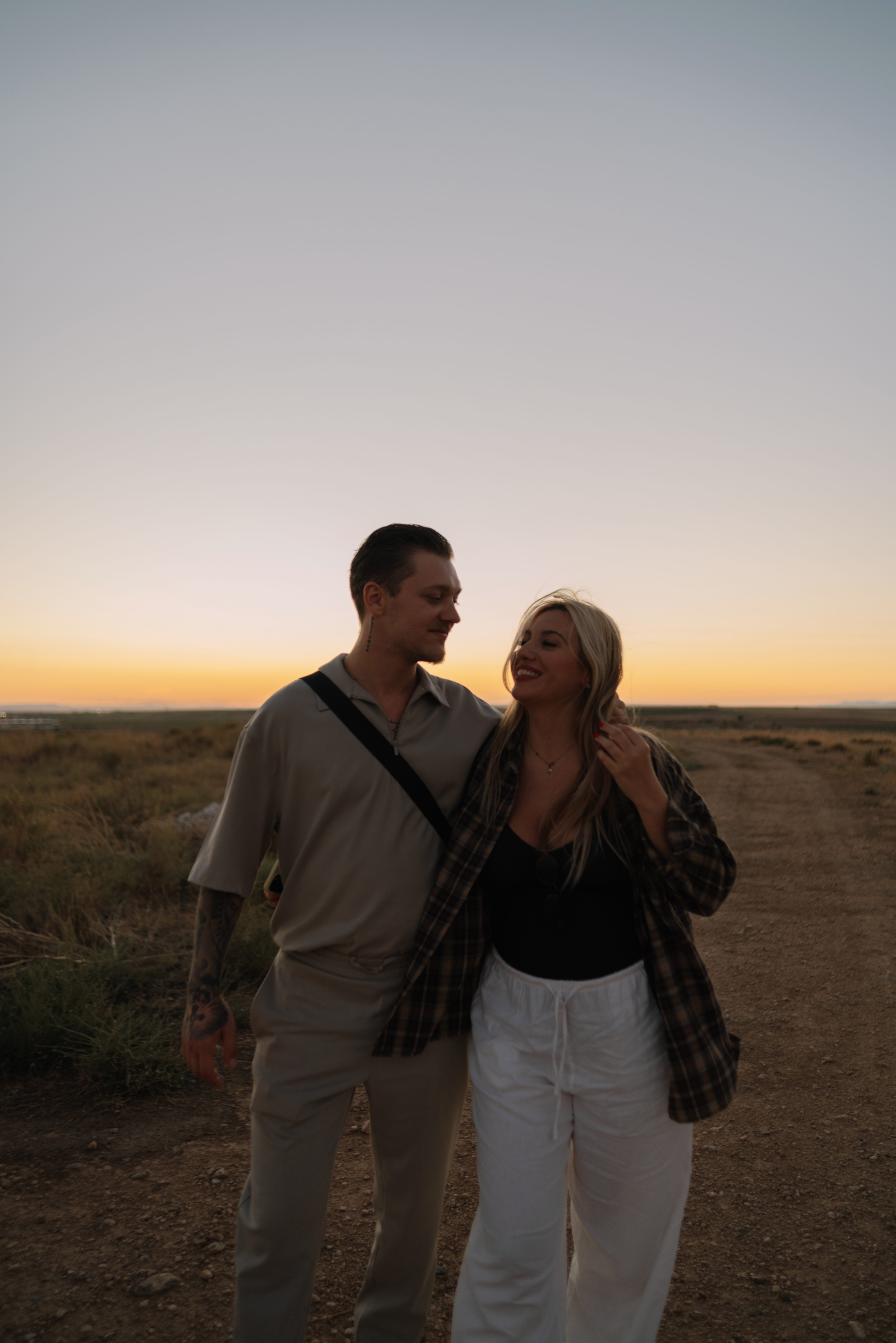 A smiling couple walks arm in arm on a dirt road at sunset, open fields and a glowing horizon behind them.