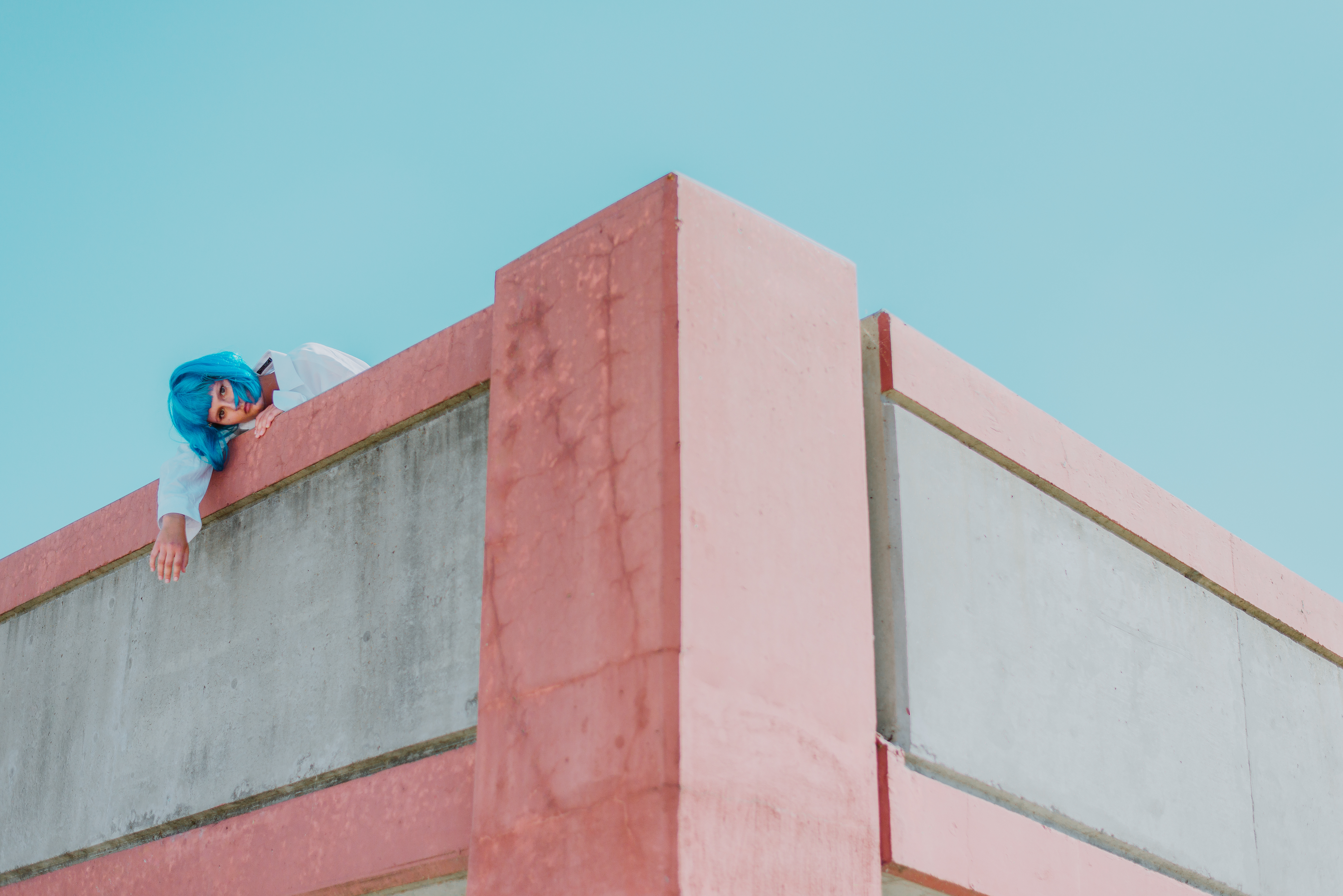 Person with blue hair leaning over a pink concrete rooftop against a pale blue sky, cool pastel color palette creating calm and distance.