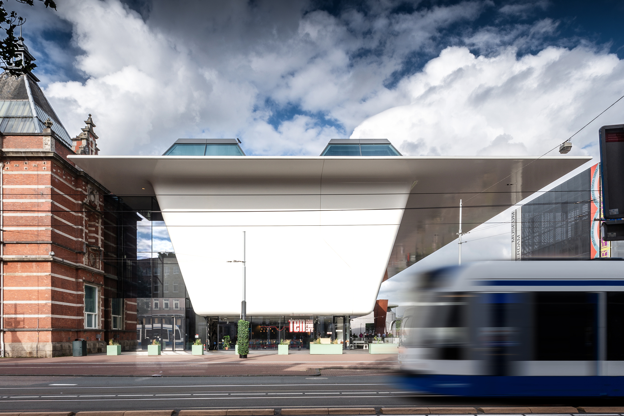 White cantilevered extension of the Stedelijk Museum in Amsterdam beside its historic brick wing, a tram blurring past in Andrew Shenouda's architectural photography.