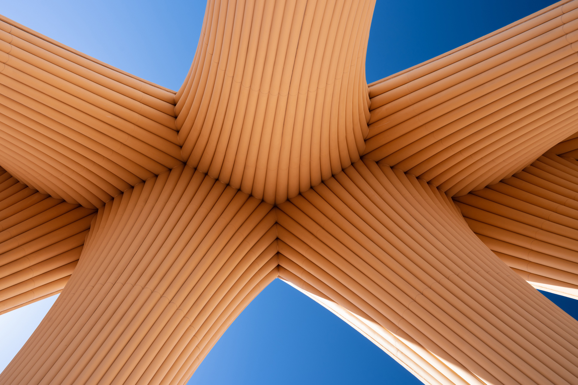 Terracotta ribbed arches meeting overhead in an X against a deep blue sky, a structural detail from Andrew Shenouda's architectural photography.