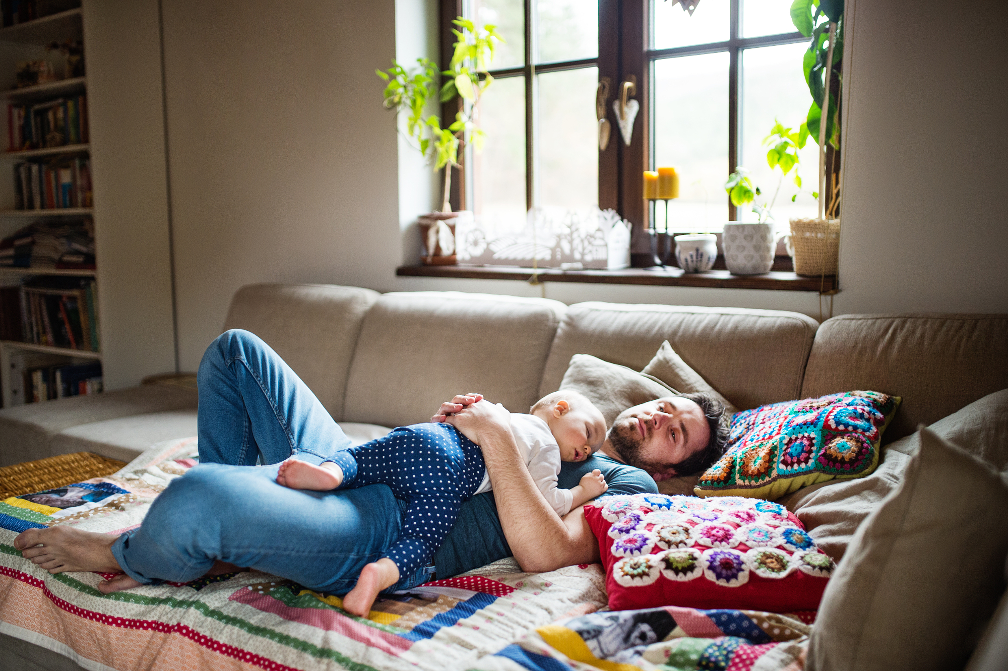 Father rests on a sofa with his baby asleep on his chest, an authentic portrait in soft natural window light.