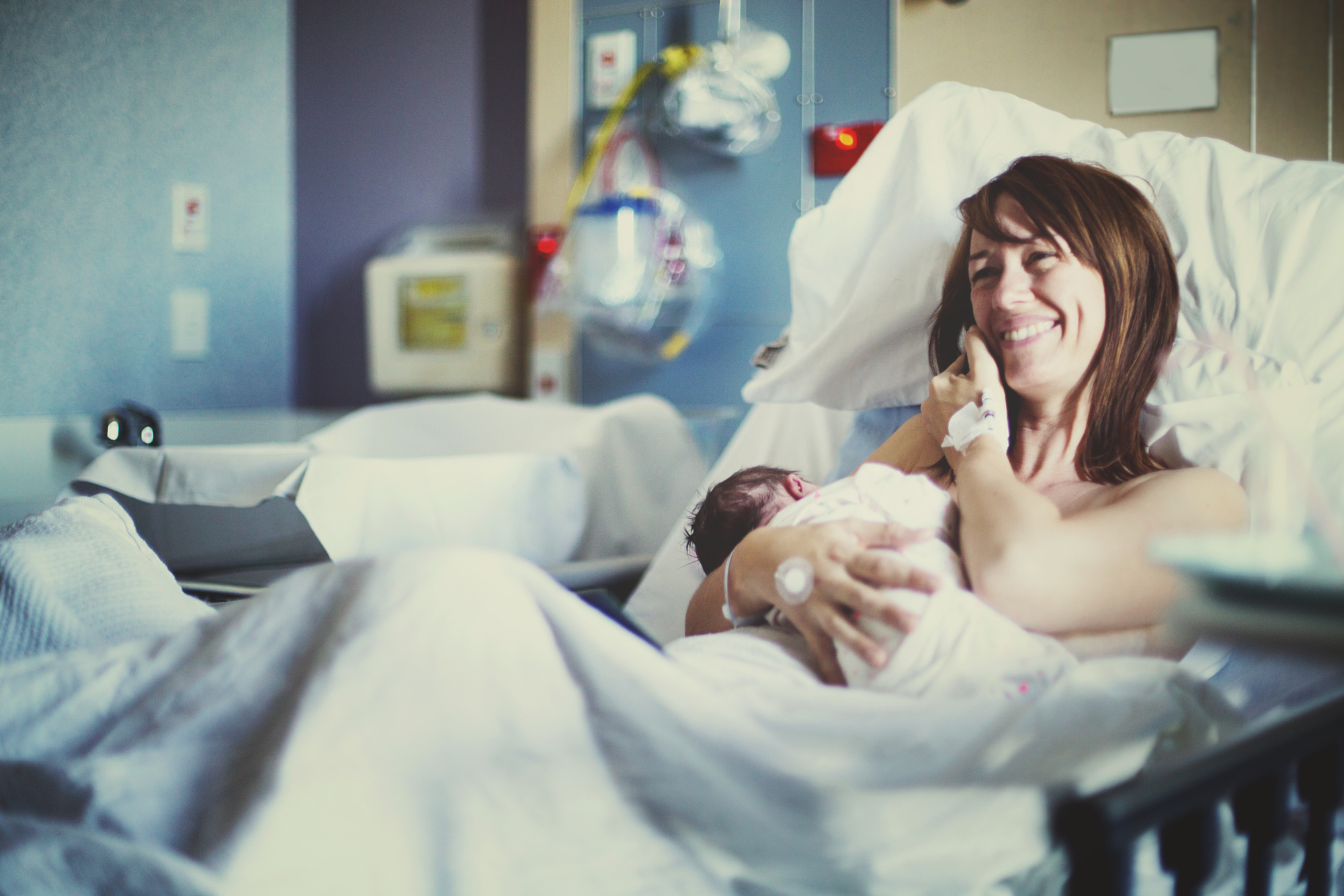 New mother smiles while holding her newborn in a hospital bed, documentary photography of a first family moment.
