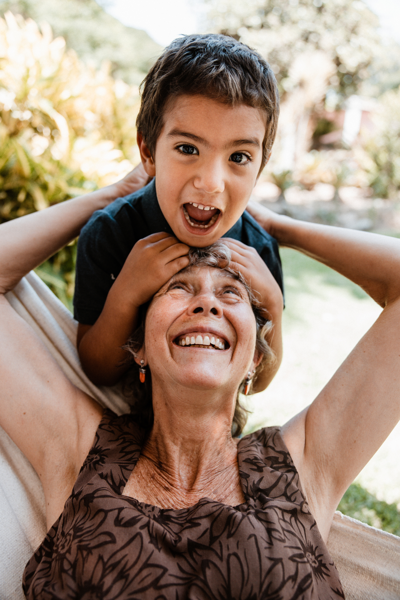 Young boy laughs on his grandmother's shoulders in a hammock, a candid photo of unguarded joy between generations.