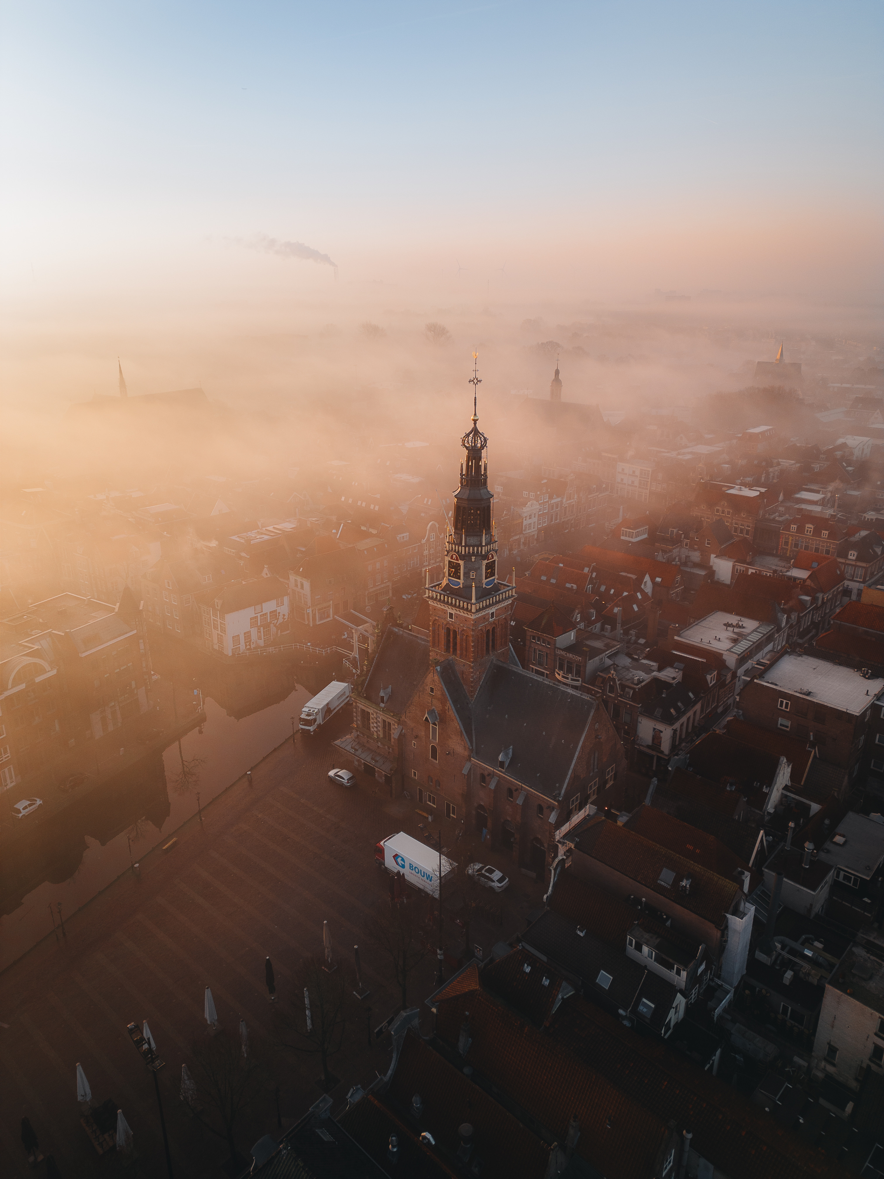 Drone view of a historic clock tower rising above a Dutch town blanketed in golden morning fog at sunrise, with canals and rooftops fading into the mist.