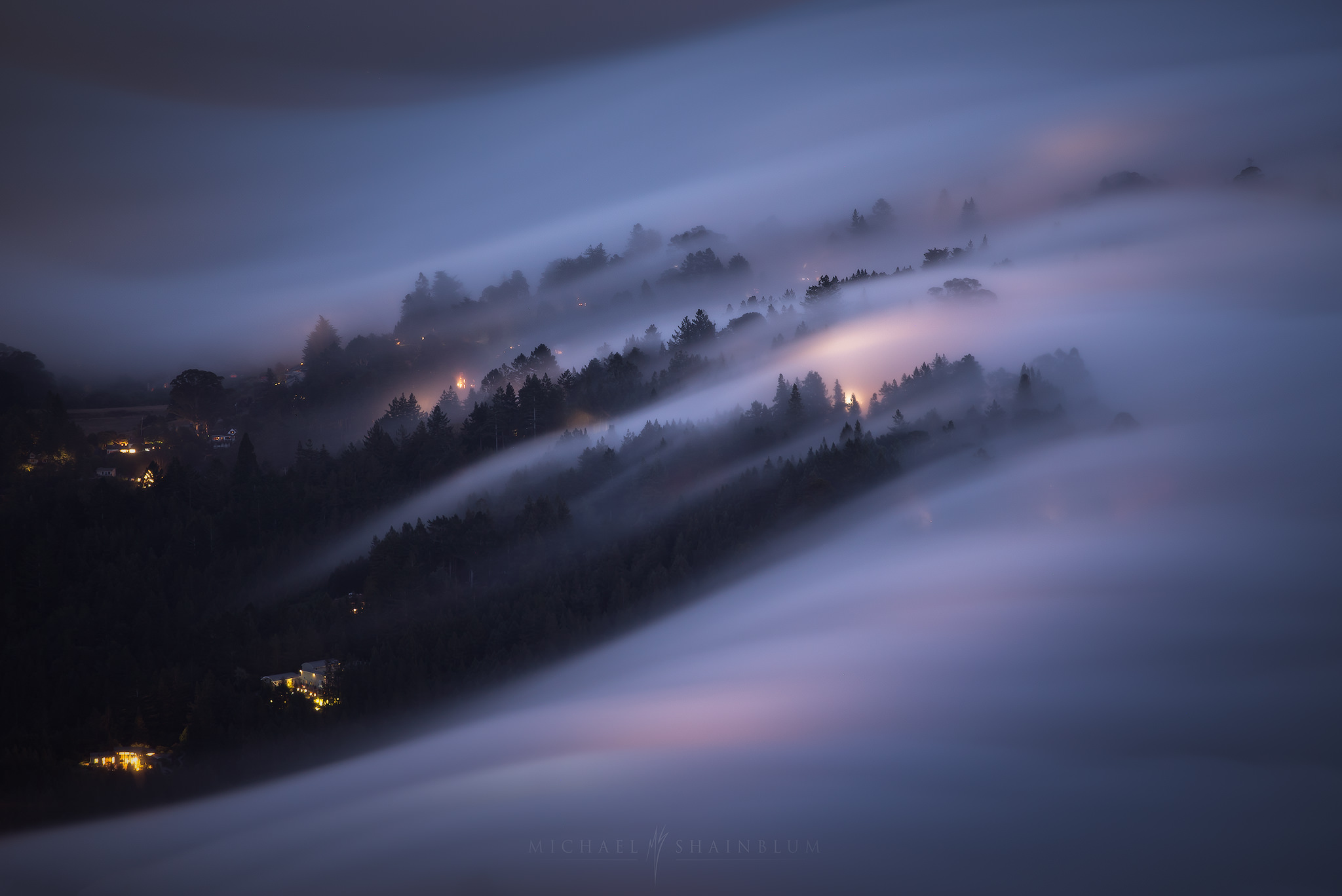 Long exposure of fog flowing over forested hills at blue hour, with layered trees and warm house lights showing atmospheric perspective in a foggy landscape.